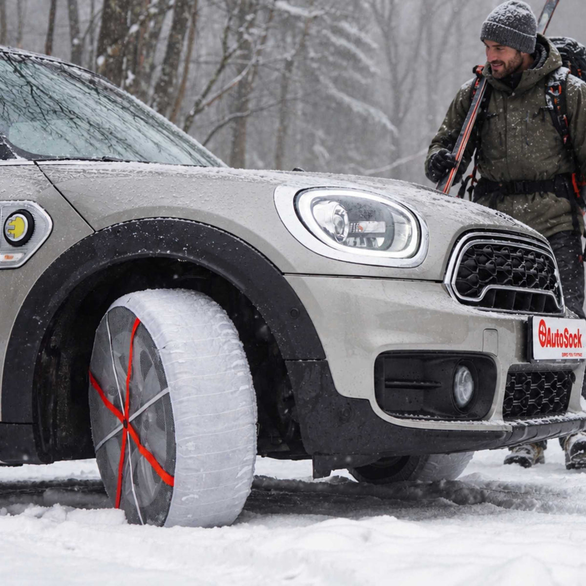 A silver Mini Cooper in a snowy outdoor setting, showing snow socks installed on the front wheel. A man in winter gear and ski boots walks towards the car and smiles.