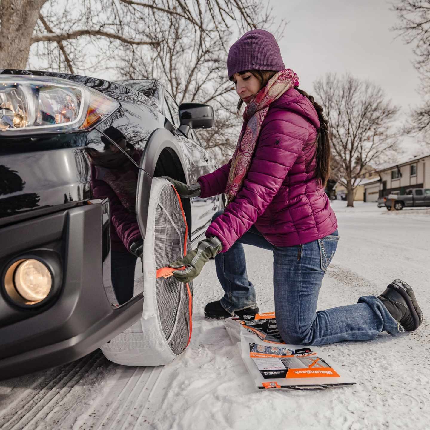 A woman mounts snow socks on the front wheel of a car in the city