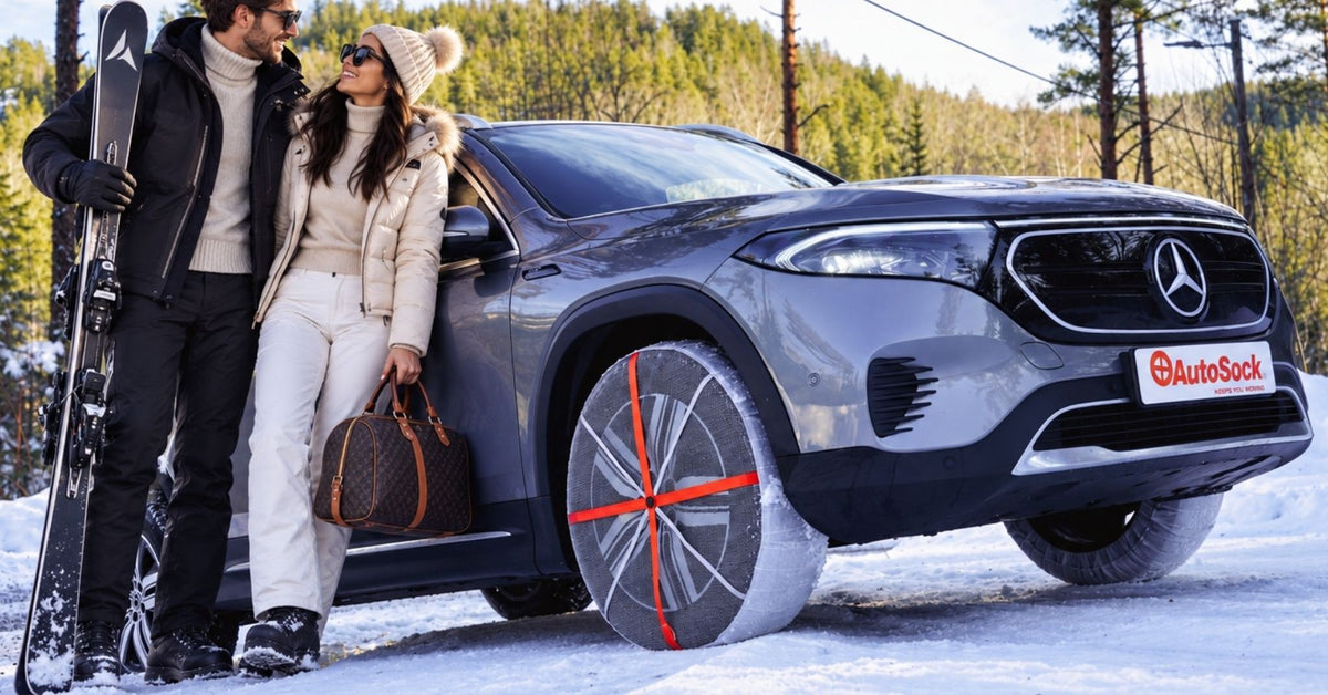 A gray Mercedes-Benz SUV with snow socks installed on winter tires, parked in a snowy outdoor setting with a couple standing beside it, one holding skis and the other holding a brown handbag. 